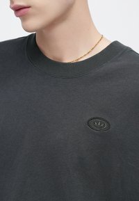 Sötétszürke pulóver kerek nyakkivágással, bal mellrészen fekete, hímzett logóval dombornyomott mintával. Sima anyag, hétköznapi dizájn.