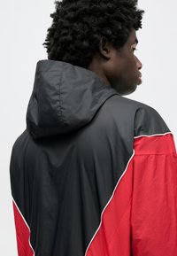 Κουκούλα με μαύρο πάνω μέρος και κόκκινο κάτω τμήμα, με αντιθετική λευκή επένδυση. Ελαφρύ υλικό με χαλαρή εφαρμογή.