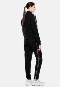 Chándal deportivo negro con chaqueta de cremallera y pantalones. Cuenta con un panel lateral brillante con logotipos, puños acanalados y una textura suave.