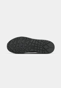 Semelle de chaussure noire avec des motifs géométriques de la bande de roulement, incluant des triangles et des rectangles, affichée à plat sur un fond blanc.