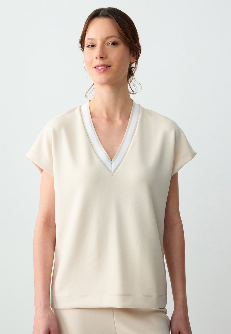 Beige short-sleeve top met een V-hals en witte rand, met een gladde textuur en een relaxte pasvorm die tot de heupen valt.