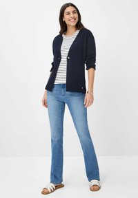 Marineblauwe cardigan met gouden knopen, wit en marinegestreept shirt eronder, lichtblauwe uitlopende jeans en witte sandalen met bruine zolen.