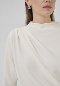 Krämfärgad topp med draperad tyg, långa ärmar och subtila veck vid halsringningen. Silverfärgade hoopörhängen syns.