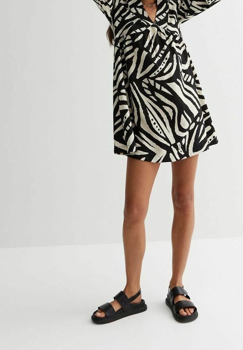 Femme portant une robe courte à motif noir et blanc et des sandales noires à bout ouvert, se tenant devant un fond clair uni.