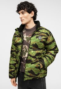 Μπουφάν puffer με πράσινο και μαύρο μοτίβο καμουφλάζ, με φερμουάρ μπροστά, ψηλό γιακά και ταμπελάκι με λογότυπο στο στήθος.