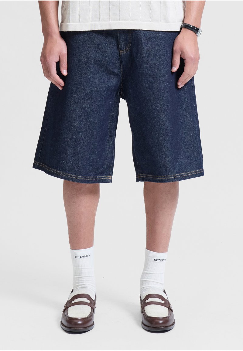 Personne portant un short en denim bleu foncé jusqu’aux genoux, des chaussettes blanches avec texte, des mocassins en cuir marron et une chemise blanche à manches courtes.