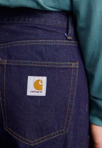 Mörkblå jeans i denim med gul sömnad och en vit etikett med en gul logotyp och text på den högra bakfickan.