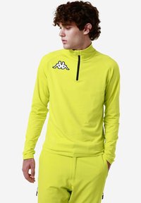 Maglietta atletica a maniche lunghe di un brillante giallo neon con colletto alto, zip a metà e logo Kappa nero sul petto. Tessuto elastico e liscio.