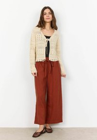 Neulecardigan vaaleassa värissä, jossa on nauha edessä, yhdistettynä löysiin, ruosteenvärisiin pellavahousuihin. Mallilla on päällään litteät, punottu ruskeat kengät.