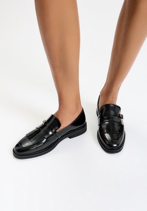 Beine, die glänzende schwarze Lederloafers mit Fransen und Schnalle auf weißem Hintergrund tragen, Füße leicht auseinander.
