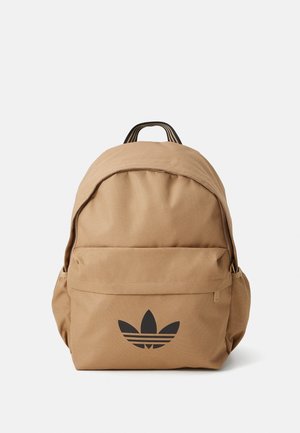 Jasnobrązowy plecak z czarnym logo Adidas trefoil na przedniej kieszeni i czarnym paskowanym uchwytem do noszenia na górze.