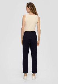 Haut sans manches beige associé à un pantalon bleu marine foncé. Le pantalon possède des ourlets retournés et deux poches arrière ; le modèle porte des chaussures claires.