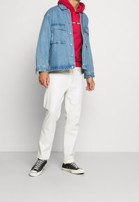 Chaqueta de mezclilla en azul claro, sudadera roja por debajo, pantalones blancos, zapatillas negras. La chaqueta cuenta con bolsillos y cierre de botón.