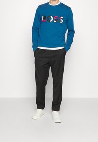 Blå sweatshirt med flerfärgad "BOSS"-logotyp, tillsammans med svarta byxor och vita sneakers. Sweatshirten har rund halsringning och ribbade ärmslut.