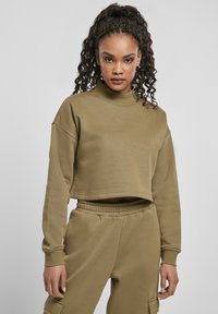 Ung kvinne med krøllete hår iført olivengrønn kortermet sweatshirt og matchende cargo-bukser, som står mot en lys, ensfarget bakgrunn.