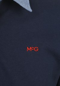 Navyblaues Strick-Polohemd mit einem hellen Denim-Kragen; mit rotem gesticktem "M@G"-Logo auf der Brust. Weicher Stoff und figurbetonter Schnitt.