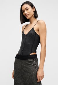 Γυναίκα με κοντά σκουρόχρωμα μαλλιά που φοράει μαύρο, ριγωτό, αμάνικο τοπ με κουμπιά και σκουρόχρωμη γκρι φούστα με μαύρη μέση.