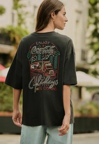 Black oversized t-shirt featuring colorful embroidered Coca-Cola design with trucks, trees, and "Enjoy Holidays, They're Coming" text.