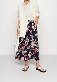 Person som bär en vit ärmlös kavaj, ljusbeige skjorta, marinblå kjol med rosa fjäril och blommönster, håller en vit handväska och har på sig vita sandaler.