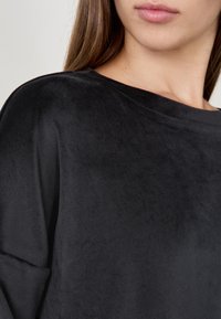 Donna che indossa un top a maniche lunghe nero, dalla texture morbida, con scollatura rotonda, che mostra la parte inferiore del viso e il collo.