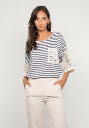 Femme portant un haut rayé gris et blanc avec des manches en dentelle et une poche, associée à un pantalon beige taille haute, debout les mains dans les poches.