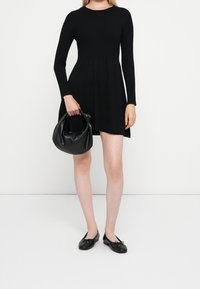 Femme portant une robe noire à manches longues, tenant un petit sac à main en cuir noir, et chaussée de ballerines noires sur un fond blanc uni.