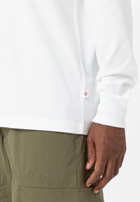 T-shirt bianco a maniche lunghe con polsini a coste, spacco laterale e una piccola etichetta con logo all'orlo. Indossato con pantaloni verde oliva con tasche cargo.
