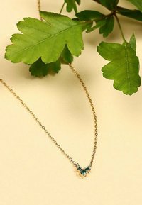 Guld kæde med en trekantet vedhæng med teal ædelsten. Set mod en lys beige baggrund og grønne blade.
