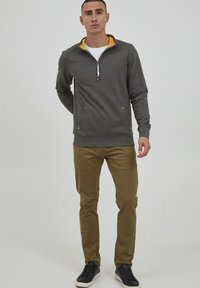 Suéter gris con un collar y cremallera amarillos, que cuenta con dos bolsillos frontales con detalles dorados. Combinado con pantalones caqui y zapatillas negras.