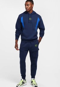 Granatowa bluza z kapturem z niebieskimi akcentami, kieszenią kangurka i sznurkiem do ściągania kaptura. Dopasowane granatowe spodnie dresowe z kieszeniami cargo. Jasne detale na butach sportowych.