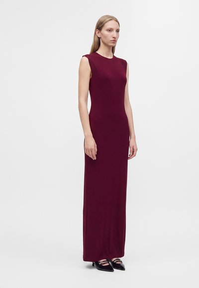 Femme debout portant une robe longue sans manches violet foncé et des chaussures noires à bout pointu sur un fond blanc uni.