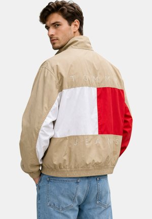 Hombre con chaqueta beige de Tommy Jeans con un gran diseño de bloques de color rojo, blanco y beige en la espalda, combinado con vaqueros azul claro.