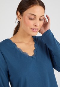 Blauwe langslijf-top met lange mouwen en een V-hals met kantafwerking. Zacht materiaal met een gladde textuur, aansluitende mouwen en een eenvoudig ontwerp.