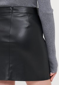 Jupe en cuir noir avec une texture lisse, taille haute et fermeture éclair au dos. Associée à un haut en tricot côtelé gris, mettant en valeur les contrastes de tissus.