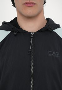 Jachetă neagră cu glugă, confecționată dintr-un material ușor, având fermoar, ajustare cu șnur și accente albastre deschise pe umeri. Logo-ul EA7 este prezent.