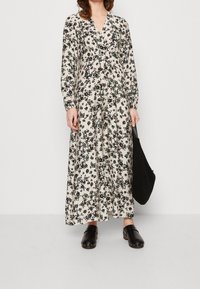 Robe maxi florale en crème avec des fleurs noires et vertes, manches longues, décolleté en V, ceinture à la taille, associée à des sabots noirs et un sac noir texturé.