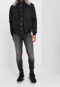 Giacca bomber nera con colletto di pelliccia grigia, indossata sopra una camicia a righe scure. Abbinata a jeans grigi consumati e sneakers nere.