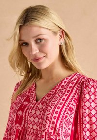 Różowa sukienka w wzory z dekoltem w kształcie litery V, z kwiatowymi i geometrycznymi motywami. Miękka tkanina o luźnym kroju i bufiastych rękawach. Srebrne kolczyki.