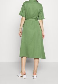 Vestito verde, lunghezza ginocchio, con maniche corte, vita legata e una texture liscia. Abbinato a sandali bianchi con tacco quadrato.