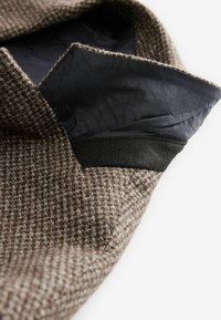 Gewebter brauner Stoff mit einem strukturierten Muster, versehen mit einem schwarzen Innenfutter und kontrastierendem Stoff am Kragenöffnung.
