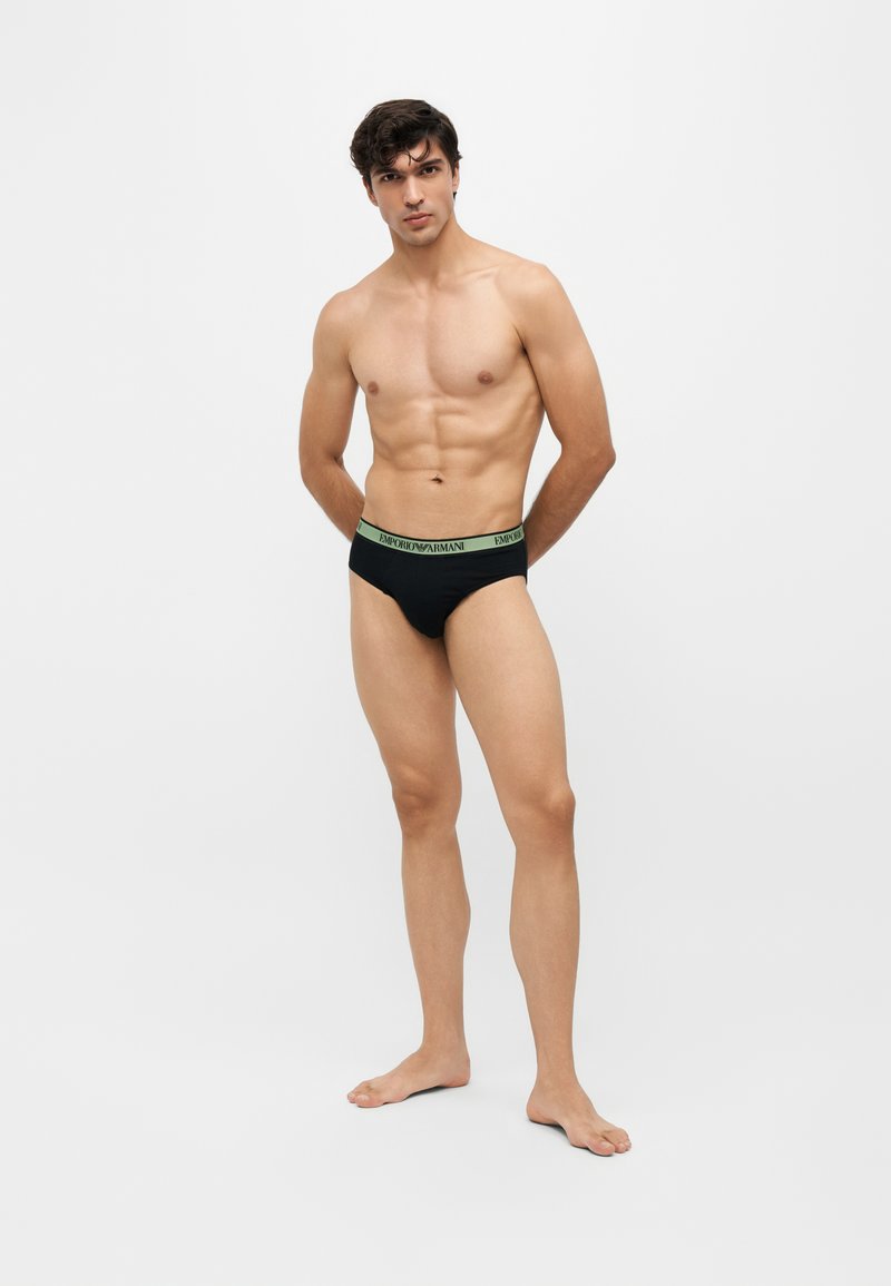 Homme musclé debout, pieds nus, portant un slip noir avec une ceinture verte et le logo Emporio Armani sur un fond clair uni.