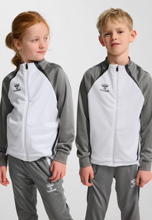 Twee kinderen in bijpassende grijs-witte Hummel sportpakken staan tegen een effen achtergrond, het meisje kijkt naar de jongen.