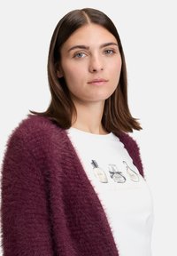 Burgundowa, puszysta kardigan nałożona na biały t-shirt z ilustracjami flakonów perfum i napisem "niezapomniany".