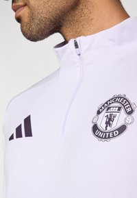 Λευκό αθλητικό πουλόβερ με φερμουάρ μέχρι τη μέση και το λογότυπο της Manchester United στο στήθος, με τρεις μαύρες ρίγες στον αριστερό ώμο. Ομαλή υφή υφάσματος.