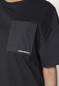Czarny bawełniany t-shirt z dużą teksturowaną kieszenią na lewej piersi, z białym haftowanym logo na dolnym brzegu. Krótkie rękawy, okrągły dekolt.
