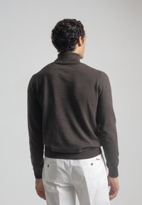 Hombre de espaldas vistiendo un suéter de cuello alto marrón oscuro y pantalones blancos frente a un fondo gris claro liso.