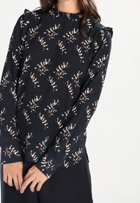 Žena oblékající tmavě modrou blůzu s dlouhým rukávem, na které jsou malé bílé květinové vzory a volánkové detaily na ramenech a výstřihu.