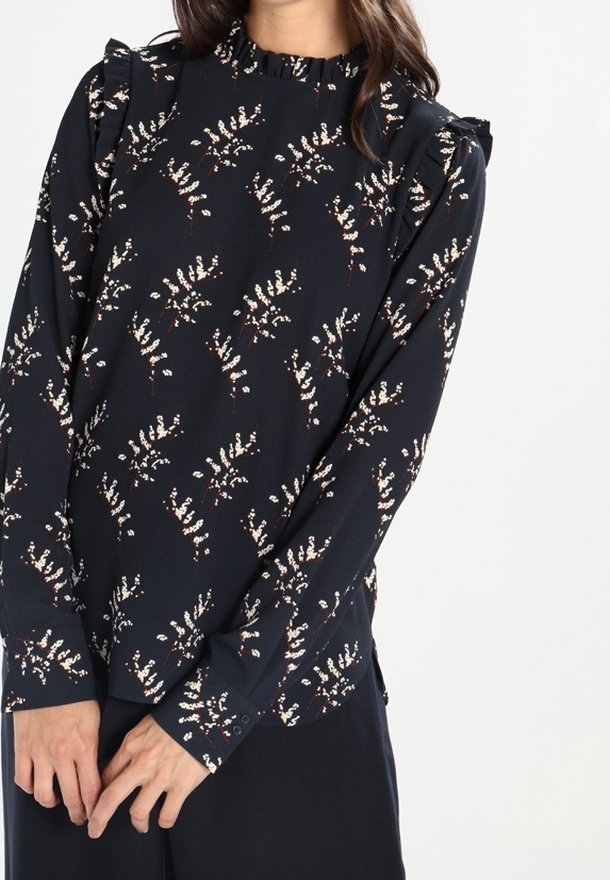 Žena oblékající tmavě modrou blůzu s dlouhým rukávem, na které jsou malé bílé květinové vzory a volánkové detaily na ramenech a výstřihu.