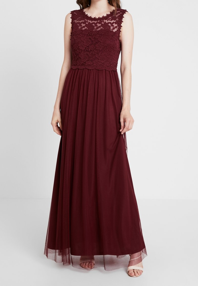 Femme portant une robe bordeaux sans manches, longue jusqu'au sol, avec un corsage en dentelle et une jupe fluide, debout devant un fond clair uni.