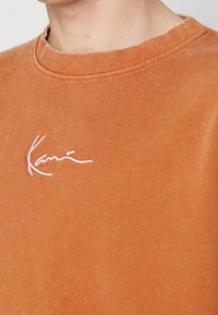 Primer plano de una persona que lleva un sudadera de cuello redondo naranja desgastada con texto en cursiva bordado en blanco en el pecho.